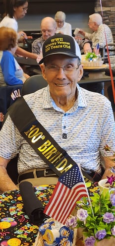 Sid Trexler celebrates his 100th birthday at Cedarhurst Senior Living, wearing a '100th birthday' sash around his chest, and a hat that says ' WWII Infantry Veteran 100 years.'