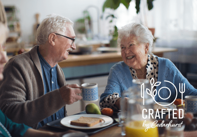 An older adult couple sit at a table enjoying their breakfast together and smiling. The man has a plate of toast and has raised his coffee mug to drink. In the bottom right corner, a white logo reads 'Crafted by Cedarhurst®'