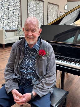 Cedarhurst of West Plains resident David Arnold sits on a piano bench. He is wearing blue slacks and a light grey jacket with a red rose pinned as a boutonniere. The community's grand piano is just behind him.