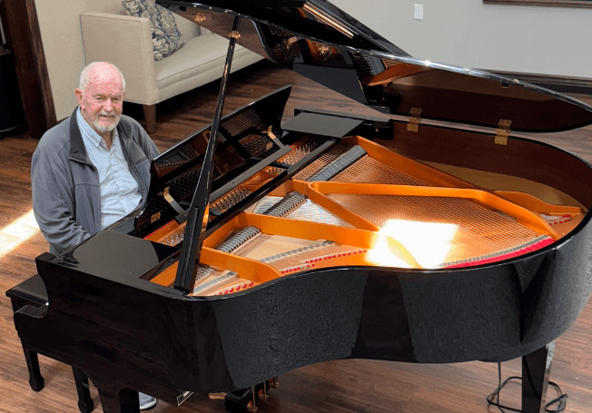 Cedarhurst Senior Living resident David Arnold sits at a black grand piano in his community. David is smiling and wearing a grey cardigan sweater and a light blue button down shirt.