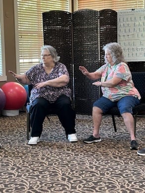 Two older adult women residents from Cedarhurst of Canton sit in an open activity room performing Tai Chi exercises in synch with one another