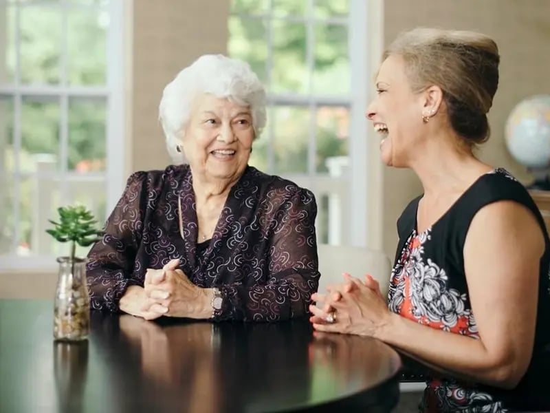 A resident and a caregiver sitting and smiling together at a dining room table