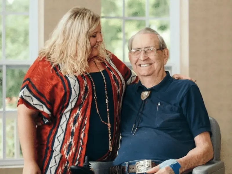 A woman with blonde hair smiles warmly at an elderly man in a wheelchair