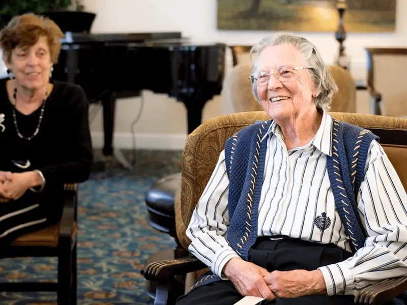 Elderly woman in striped shirt and blue vest smiling while seated in a cozy room
