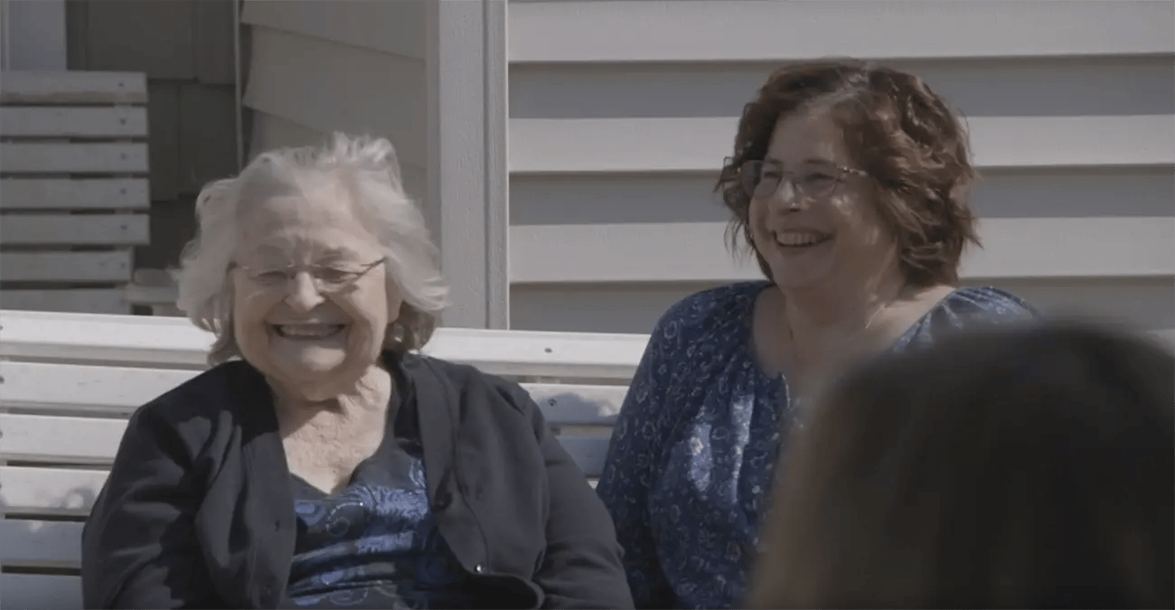 Two women are sitting on a bench outdoors, smiling and sharing a joyful moment