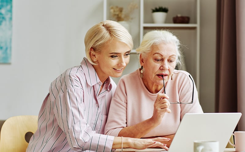 Two women are seated at a table, looking at a laptop. The younger woman is smiling, while the older woman is holding glasses, appearing focused.