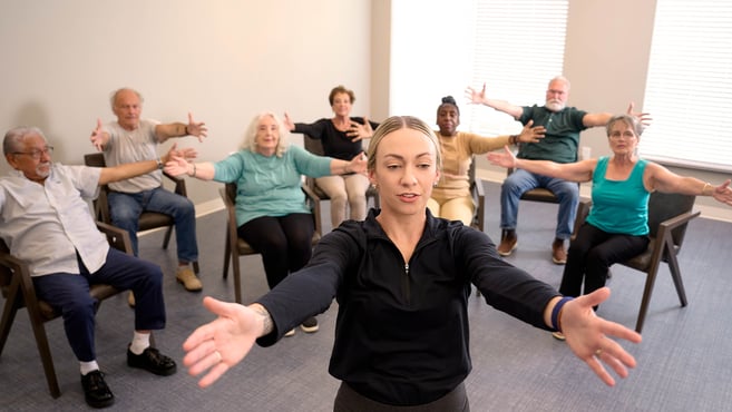 A fitness instructor leads a group of seniors in a chair exercise class at their assisted living community.