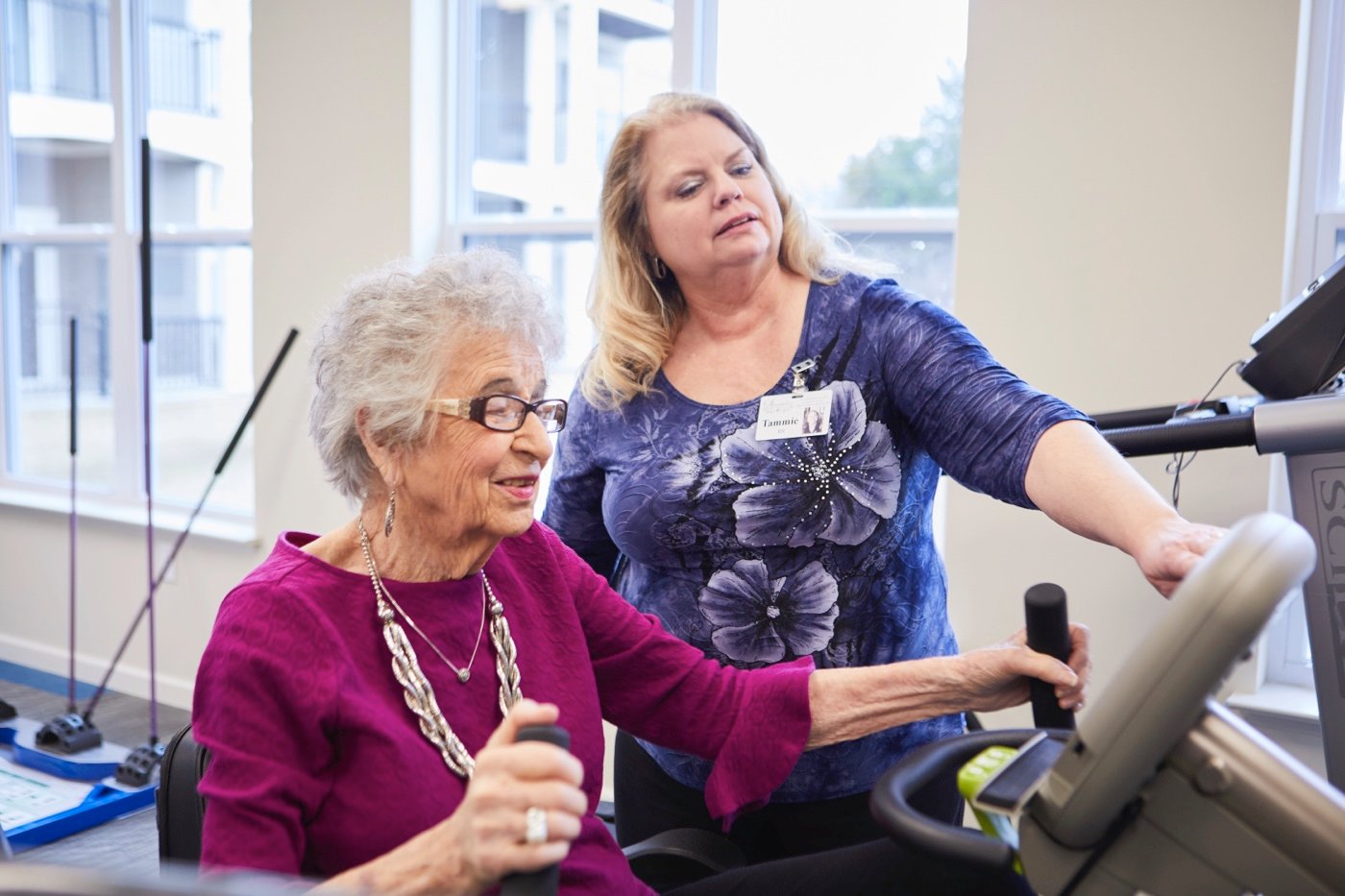 senior woman riding exercise bike with staff supervising