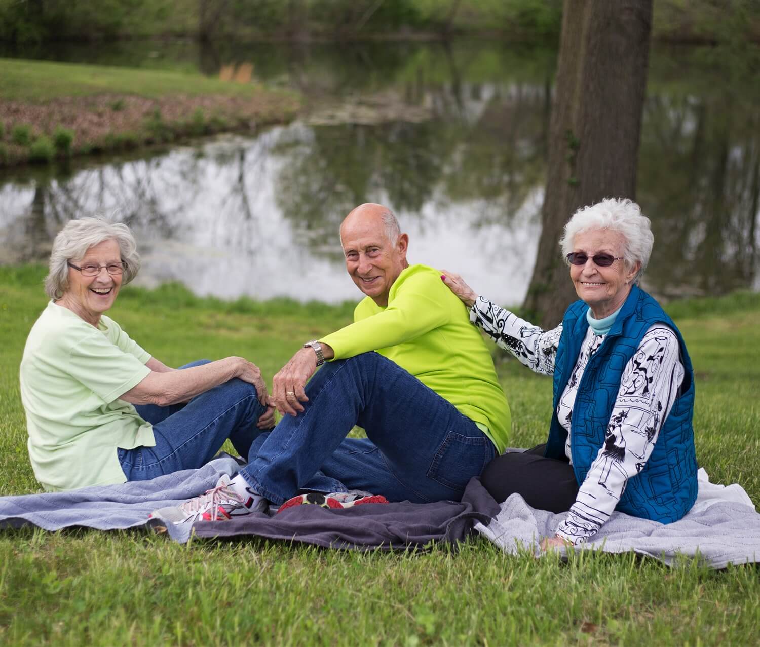 Three seniors on a picnic blanket beside a pond