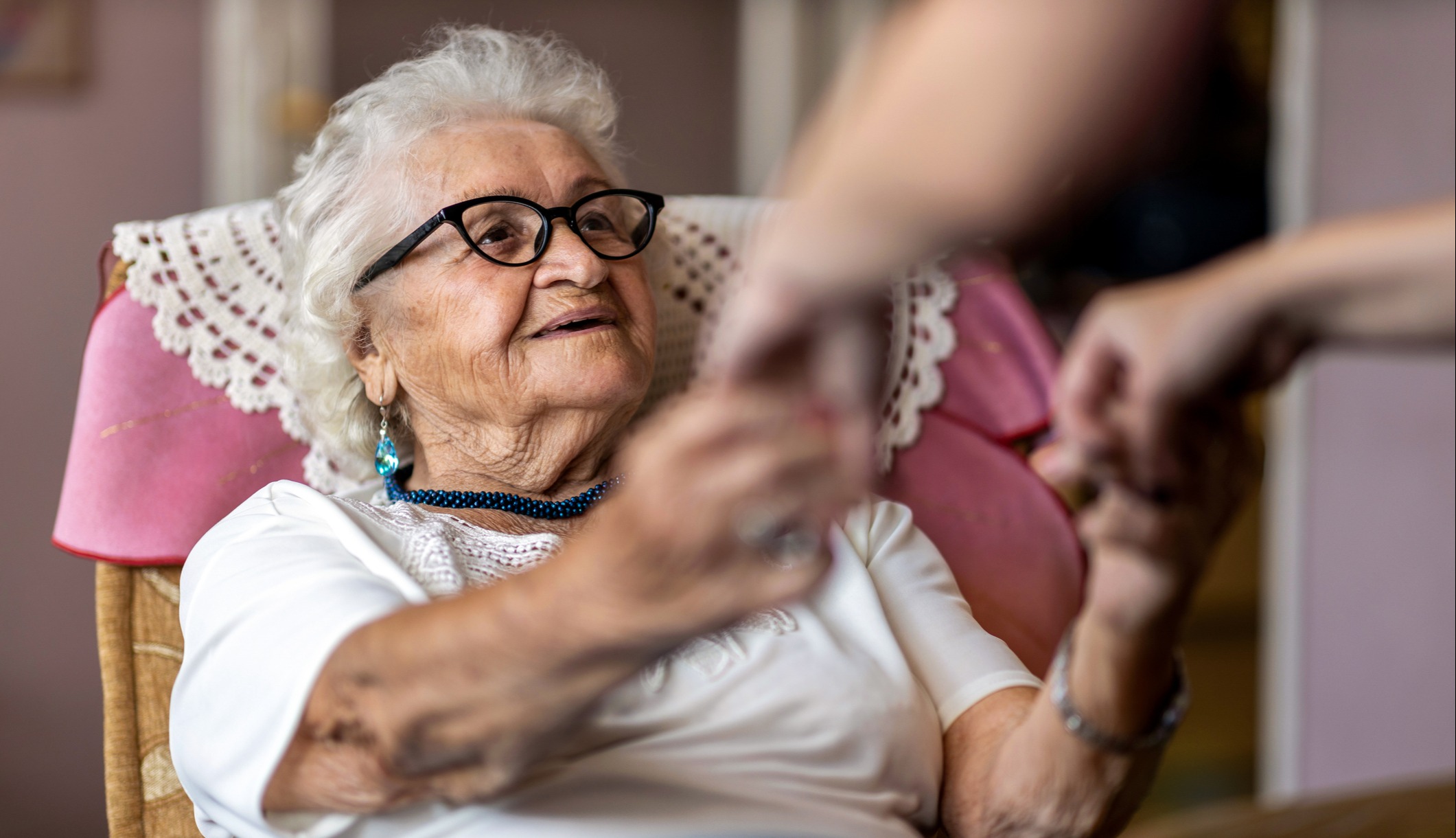 A seated senior woman gazes up at someone as they hold her hands