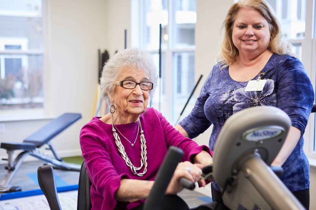 Senior resident exercising on a stationary bike as a trainer cheers her on