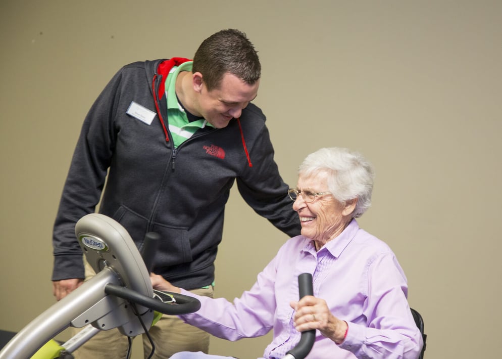 A senior resident exercising on a stationary recumbent bicycle as a trainer helps