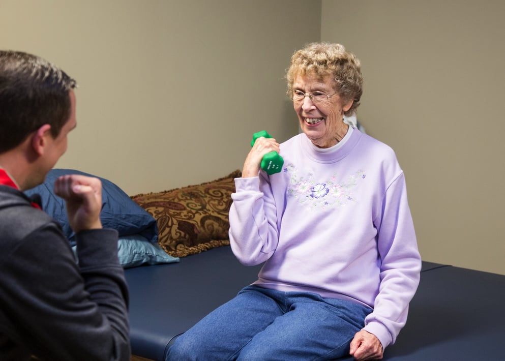 A senior resident exercising with a dumbbell as a trainer looks on