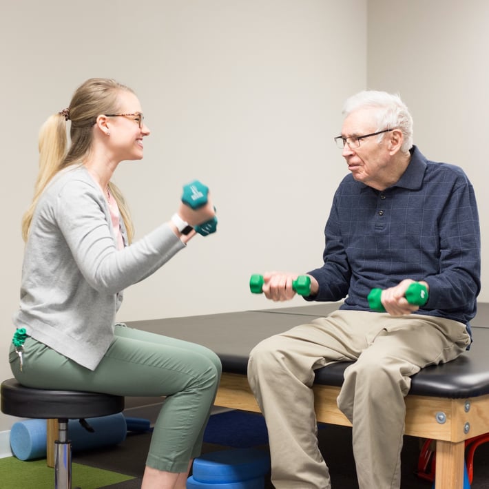 Resident in physical therapy, using hand weights with a trainer