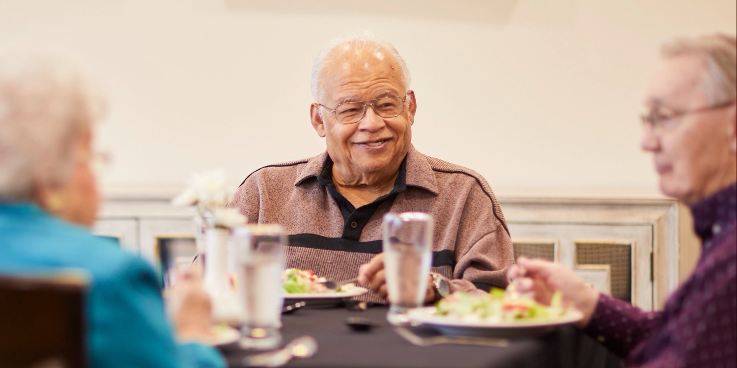 Senior man seated at a table with two friends, smiling and conversing over a meal