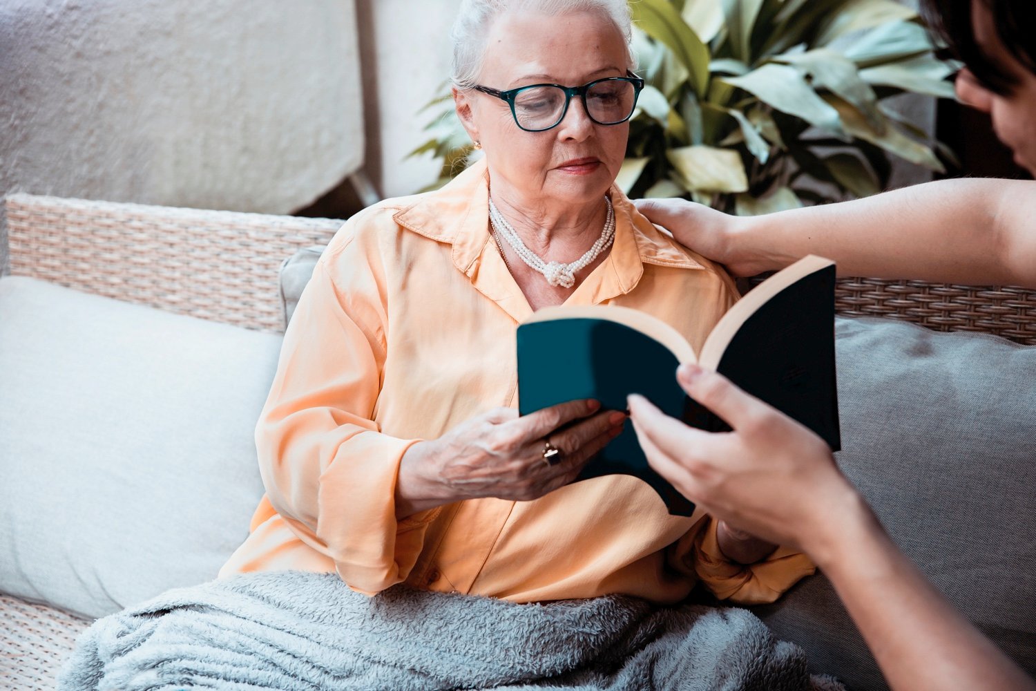 A senior woman seated on a couch, reading with the help of a staff member