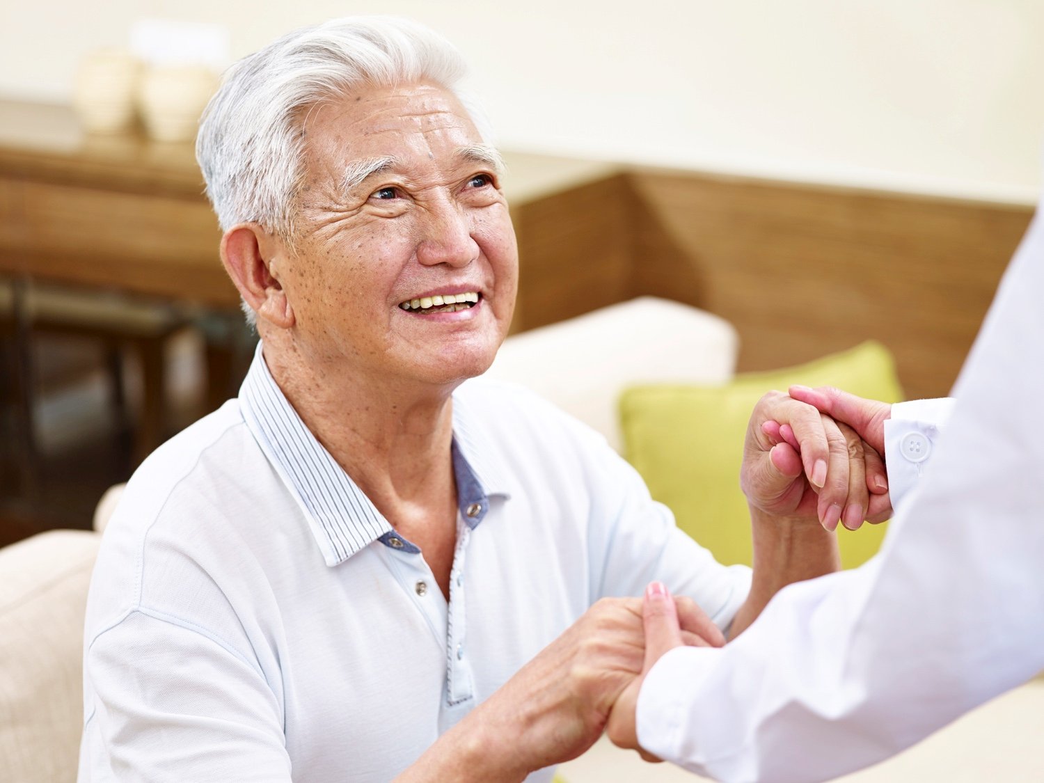 A seated senior man gazes up at someone holding his hands and ready to assist him