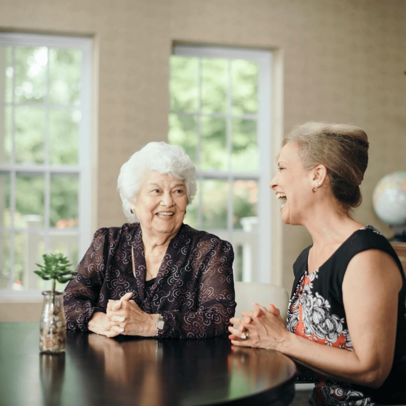 A resident and a caregiver sitting and smiling together at a dining room table