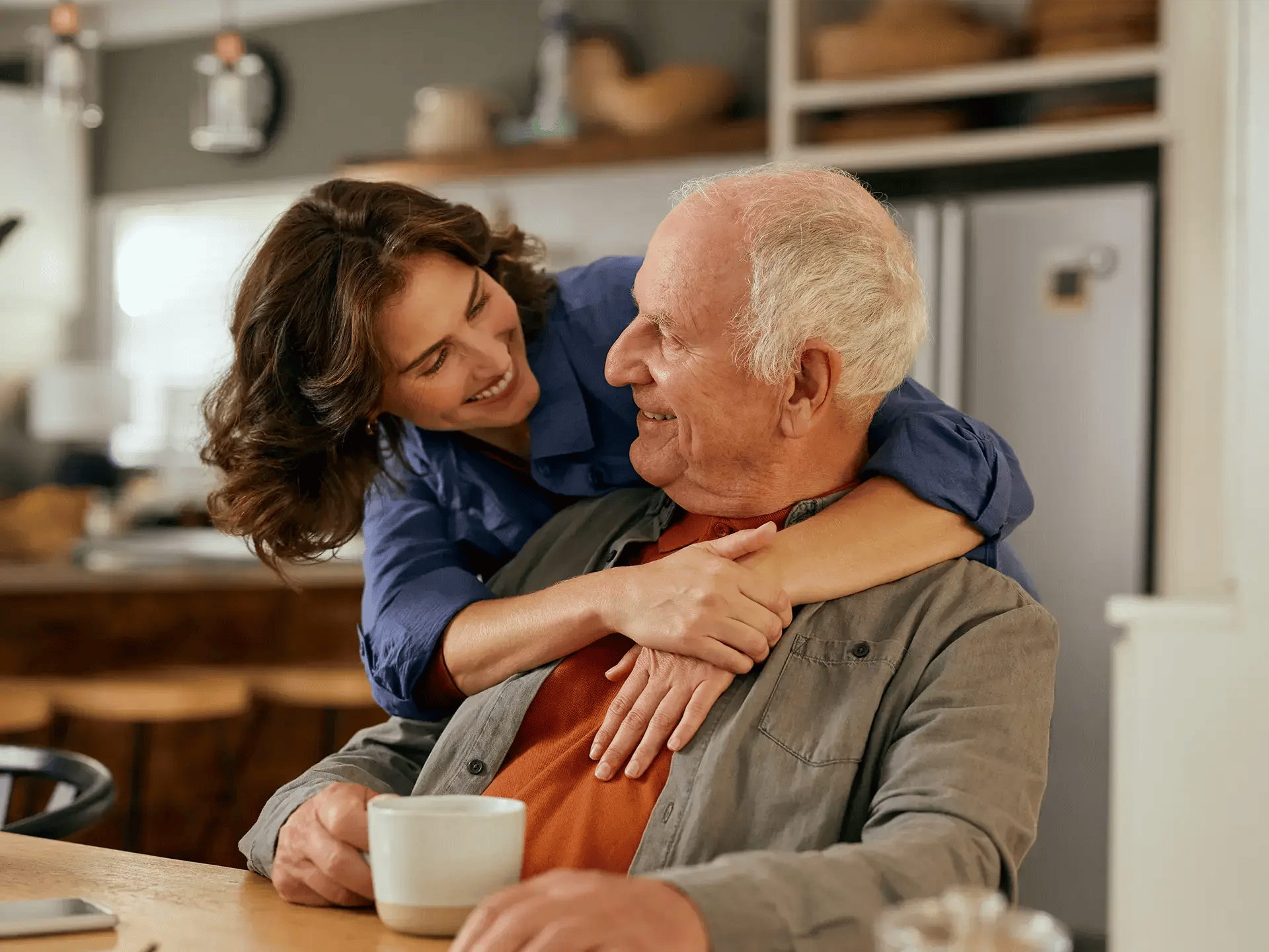 A smiling woman embraces an elderly man from behind in a cozy kitchen