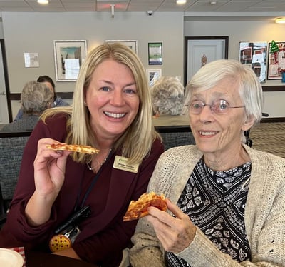 A senior living resident poses for a photo with a senior living staff member in a community common area. They are smiling and holding slices of pizza.