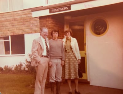 A sepia-toned photo of a three-person family — a father, mother, and young adult — posing in front of an old English home named 'Pengwern.'
