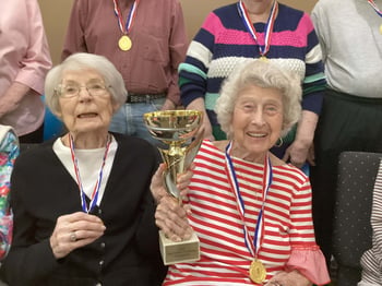 Two senior women sit together wearing gold medals around their necks and smiling. One woman, wearing a white and red horizontal striped shirt, holds a gold trophy that reads 'May Fitness Challenge'