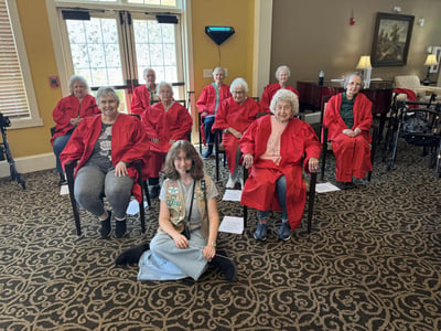 A choir club of older adult women sit and pose for a photo in red cloaks. The club leader, a girl scout, sits cross-legged on the floor in front of the club, holding a conductor's baton in her hand.