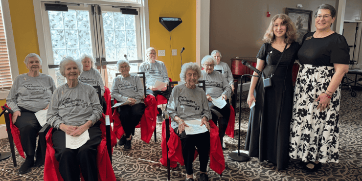 A choir club at a senior living community gathers and poses for a photo. The choir members all wear grey shirts that read 