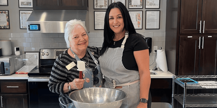 A senior living resident and a senior living staff member stand in a kitchen both wearing aprons. They both smile while standing over a large mixing bowl.