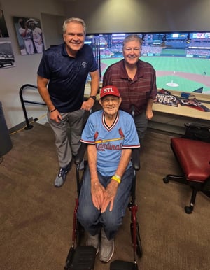 A senior living resident sits in a broadcast booth at Busch stadium alongside two KMOX sportscasters.