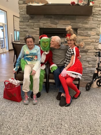 A senior couple poses for a photo at a Holiday Party at their senior living community. A person dressed as the grinch is seated between the man and woman. The man playfully leans in to give the Grinch a kiss on the cheek.