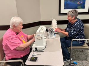 Two senior women sit at a table and use a sewing machine.