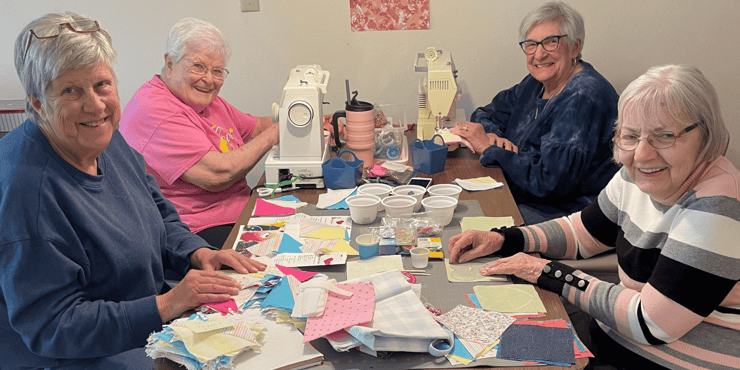 Four senior women gather around a crafting table topped with a sewing machine and many fabric swatches.