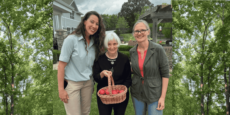 A senior woman holds a wicker basket of apples and smiles, posing for a photo with two women: the Life Enrichment Coordinator of her senior living community and the Head of Education at her community's local Forest Preserve District.