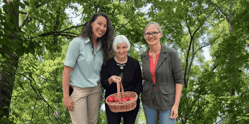 A senior woman holds a wicker basket of apples and smiles, posing for a photo with two women: the Life Enrichment Coordinator of her senior living community and the Head of Education at her community's local Forest Preserve District.