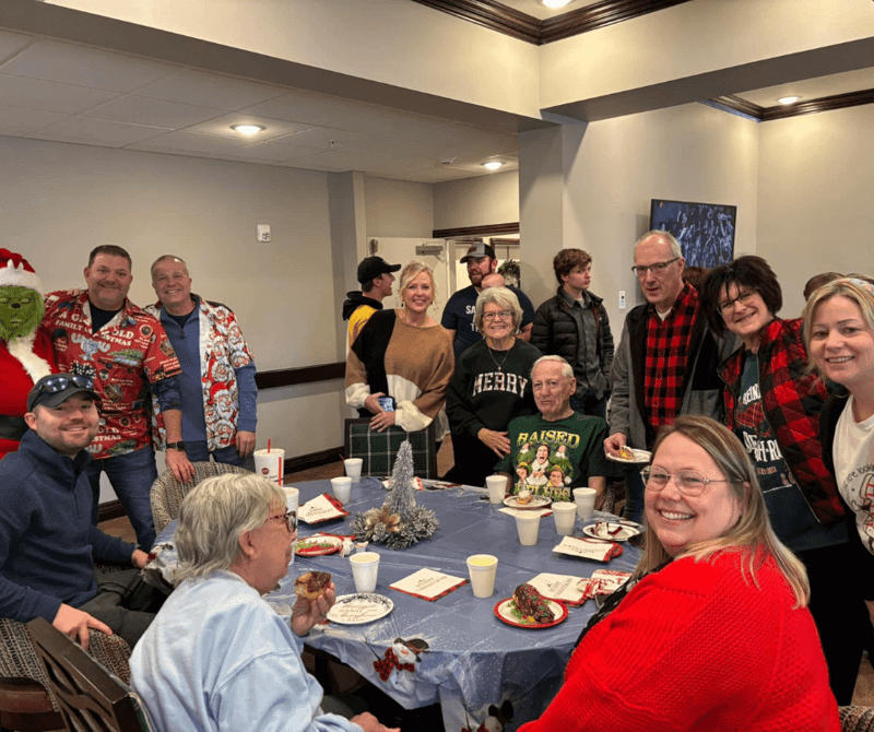 Holidays at Cedarhurst - Senior living residents and their families sit around a table, smiling and enjoying festive treats like Christmas-themed donuts. At the edge of the frame, the Grinch Who Stole Christmas poses with a smiling community staff members.