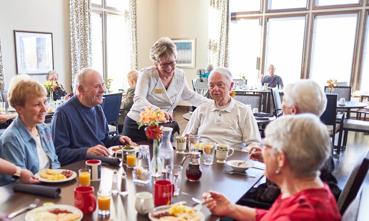 A staff member serving coffee to a group of six senior residents at a table in the community dining room