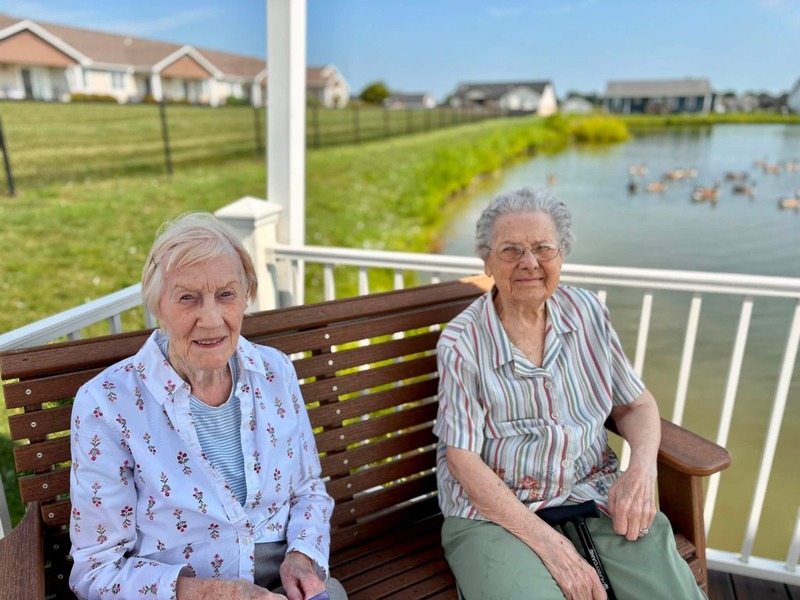 Two friends relaxing on a porch swing in a gazebo overlooking the pond at Cedarhurst of Bethalto senior living community
