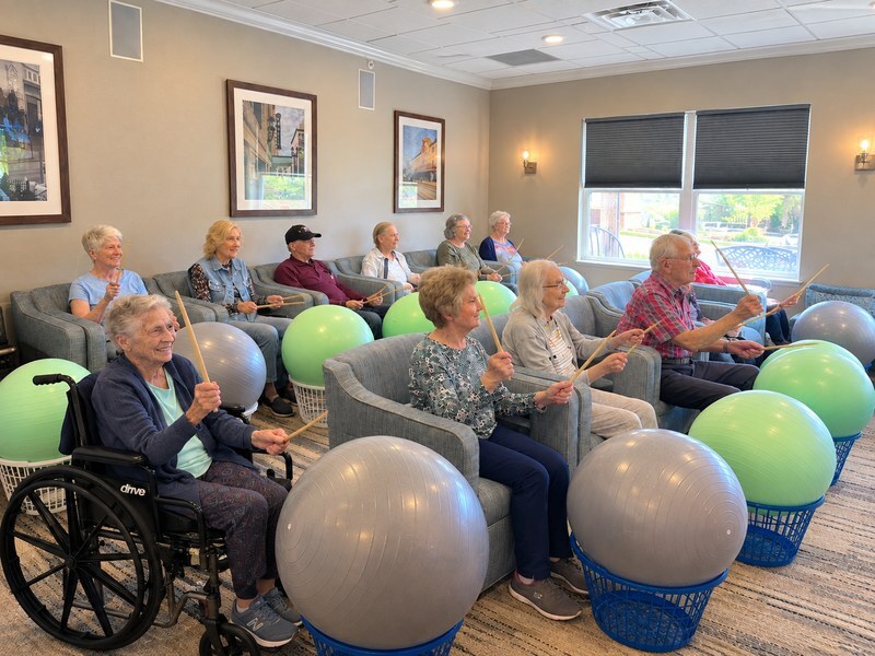 Assisted living residents taking a drum fitness class with exercise balls and drumsticks.