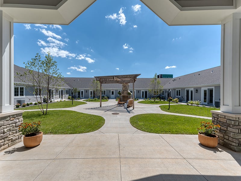 An inviting outdoor area at Cedarhurst of Farmington, showing lush green spaces, paved paths, raised garden beds, a pergola, and a covered patio