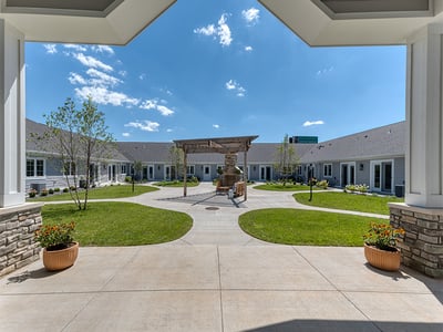 An inviting outdoor area at Cedarhurst of Farmington, showing lush green spaces, paved paths, raised garden beds, a pergola, and a covered patio