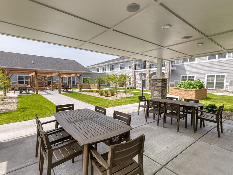 An inviting outdoor area at Cedarhurst of McHenry, showing lush green spaces, paved paths, raised garden beds, a pergola, and a covered patio.