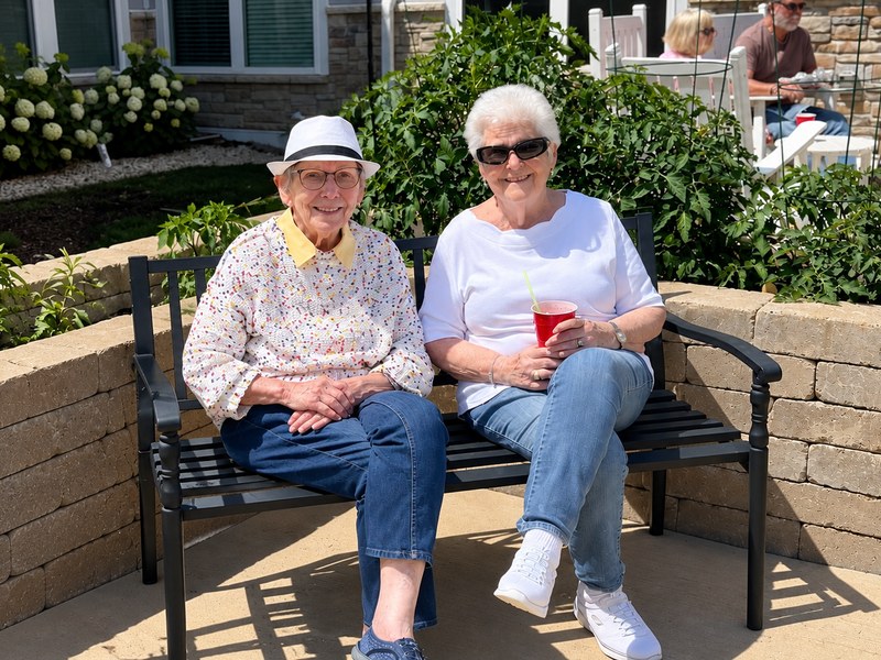 Two senior living residents smiling together on a garden bench on a sunny patio.