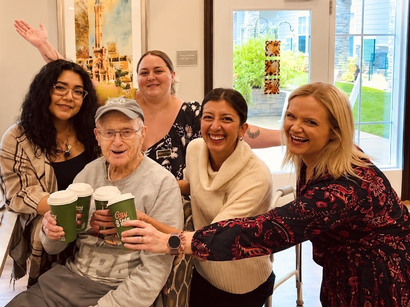 Senior living resident sharing a coffee toast with team members in a bright common area.