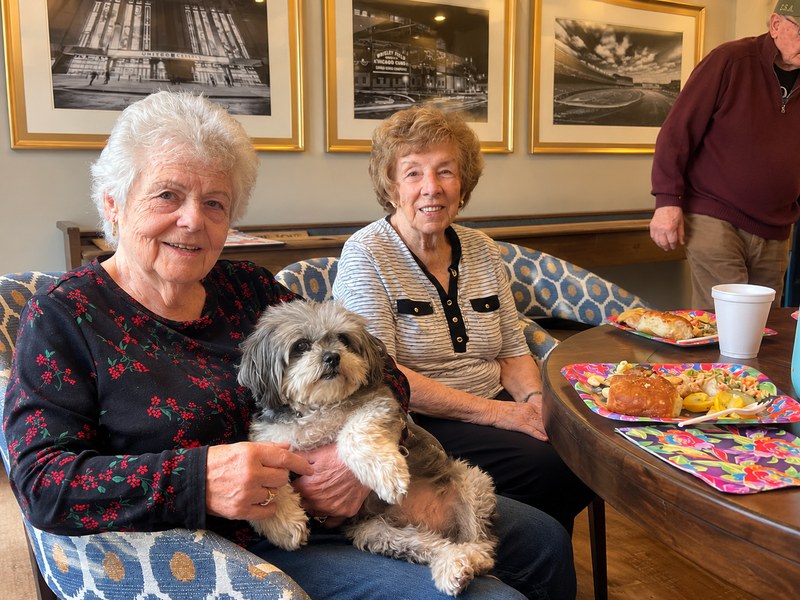 Senior living residents sharing lunch with a small dog in a comfortable common area.
