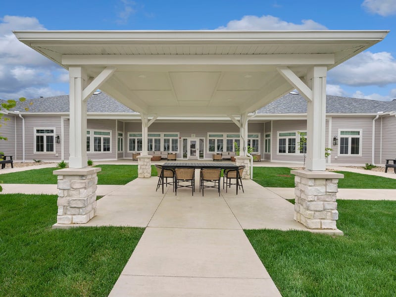 An inviting outdoor area at Cedarhurst of Newburgh, showing lush green spaces, paved paths, a pergola, and a covered patio.