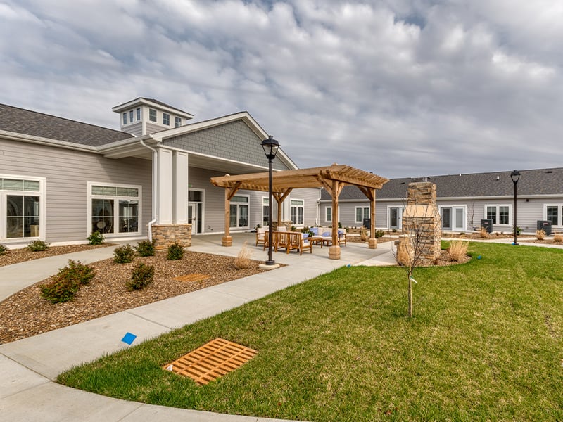 An inviting outdoor area at Cedarhurst of Owensboro, showing lush green spaces, paved paths, comfortable seating, a pergola, and a covered patio.