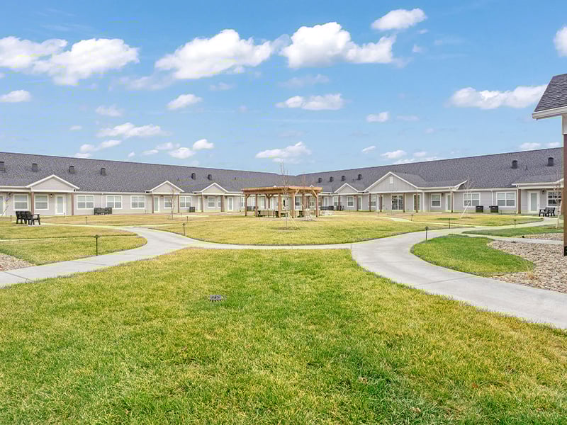 An inviting outdoor area at Cedarhurst of Salina, showing lush green spaces, paved paths, comfortable seating, a pergola, and covered patios