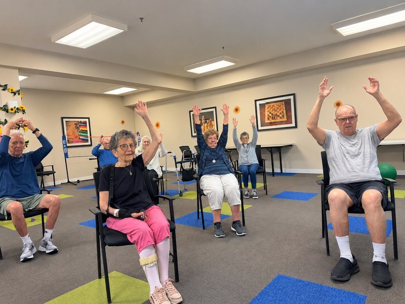Assisted living residents stretching their arms overhead during a chair exercise class.