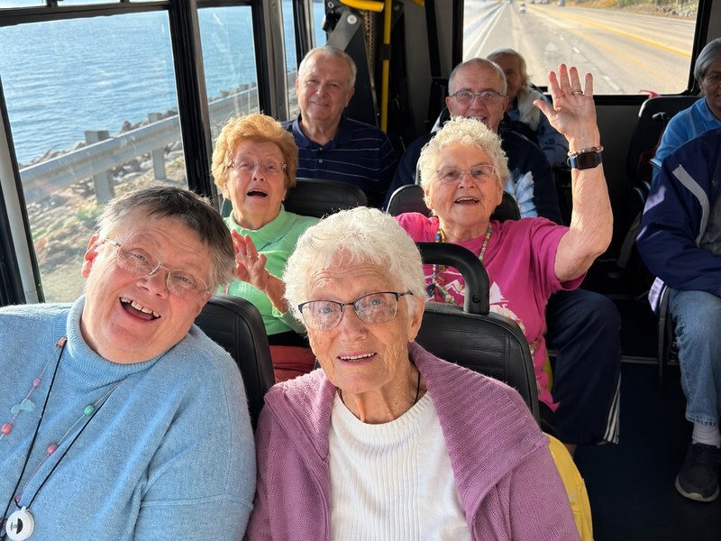 Senior living residents smiling and waving on the community bus during a scenic drive along the water